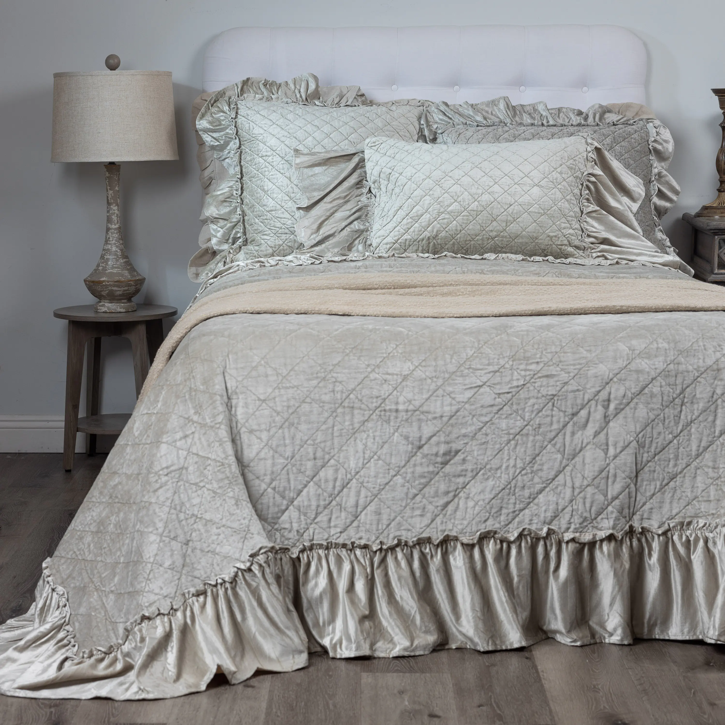 Bed with light gray quilted bedding and ruffled edges flanked by wooden nightstands and lamps on both sides of the bed
