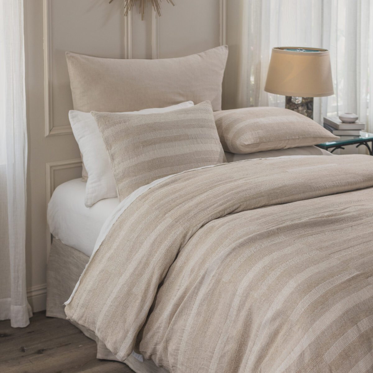 Bed with beige striped bedding and pillows in a sunlit bedroom with a lamp and stacked books on a side table It captures a cozy neutral toned sleeping nook