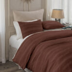 Bed with a rust colored quilt and white pillows beside a beige lampshade on a nightstand in a bright bedroom corner