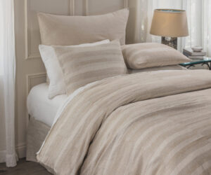 Bed with beige striped bedding and pillows in a sunlit bedroom with a lamp and stacked books on a side table It captures a cozy neutral toned sleeping nook