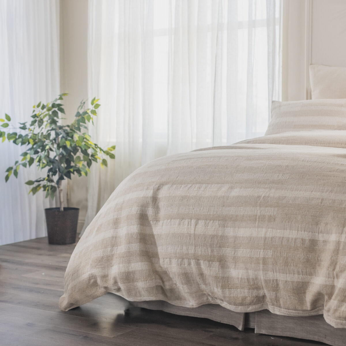 Bedroom scene with a bed covered in beige striped bedding sheer white curtains behind and a potted plant to the left