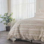 Bedroom scene with a bed covered in beige striped bedding sheer white curtains behind and a potted plant to the left