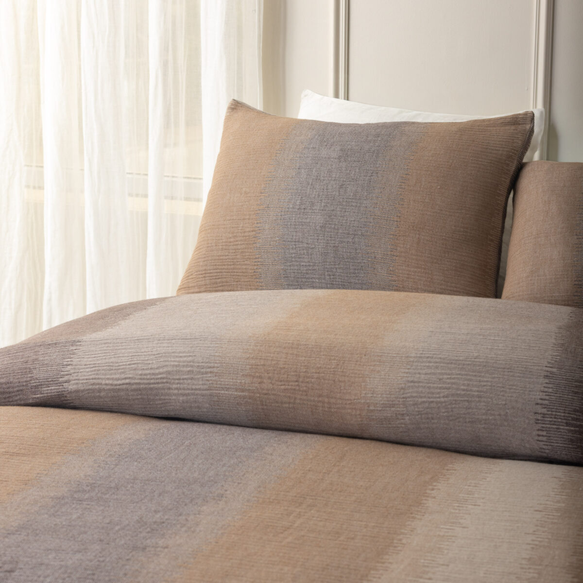 Brown and gray patterned throw pillow on a beige sofa with sheer white curtains in the background