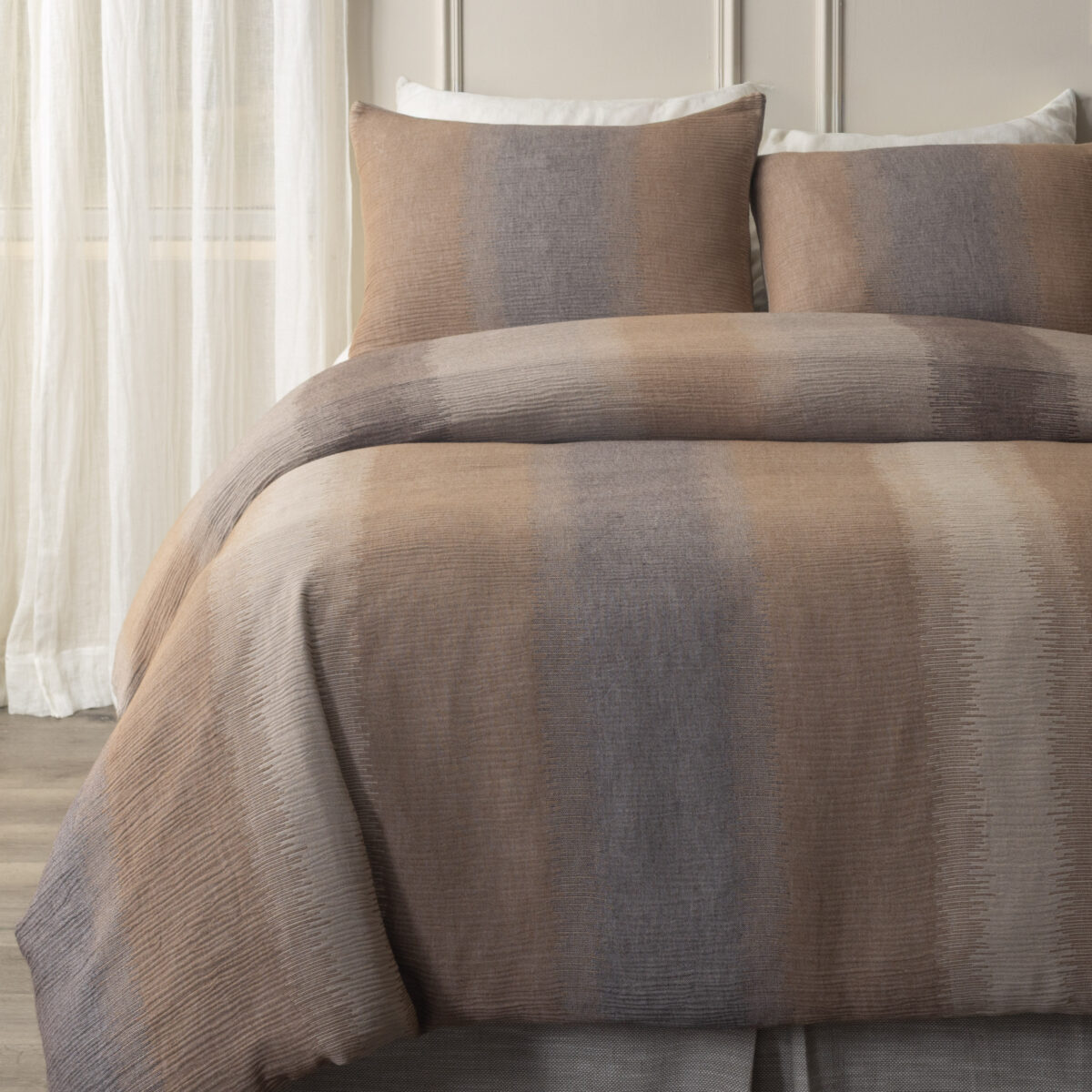 Close up of beige and gray textured bedding with two matching pillows against a headboard and light sheer curtains in the background