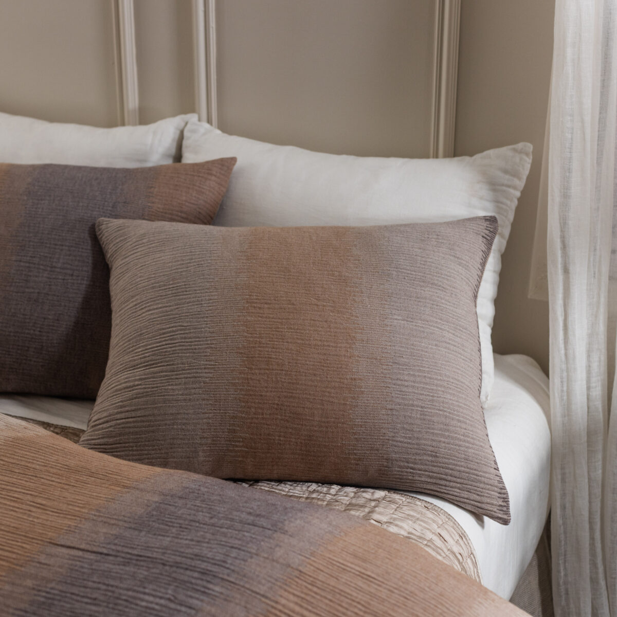 Twin layered throw pillows in neutral browns on a bed with a light headboard and sheer curtains in the background