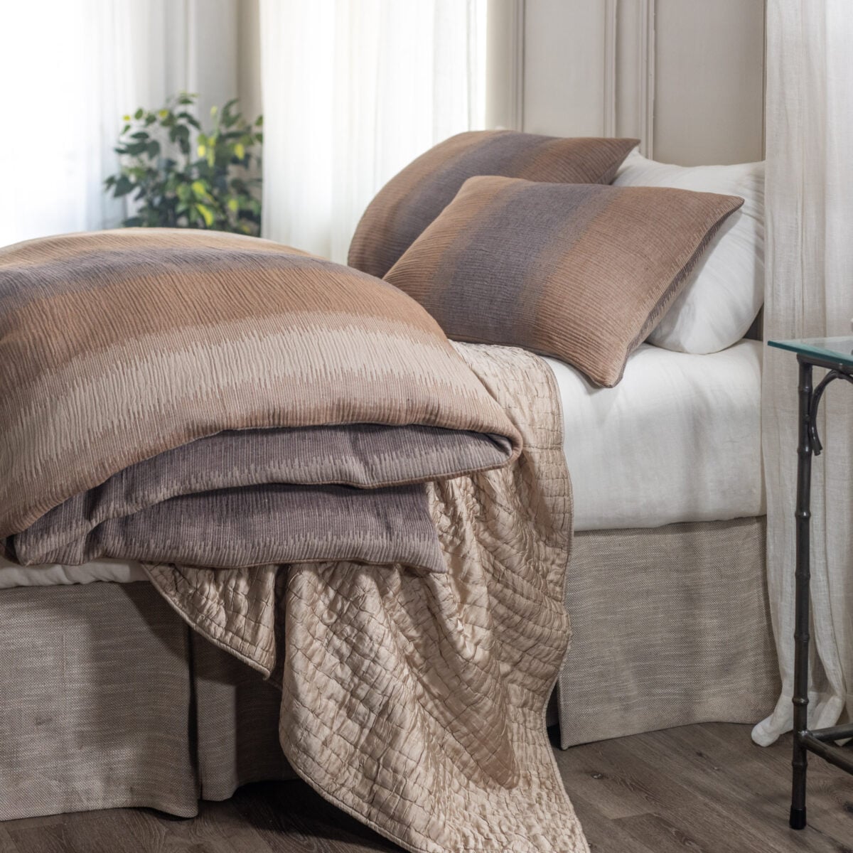 Bed with tan and brown quilted bedding and pillows in a softly lit room with white curtains in the background