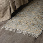 Beige quilted bedspread draped over a dark wood floor beside a light gray woven rug with a fringed edge and scattered knobbly texture