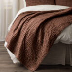 Brown quilted bedspread draped over white sheets at the foot of a bed with a wooden floor visible nearby