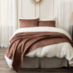 Bed with white linens and brown quilted throw pillows in front sunburst wall decor above sheer curtains framing the window