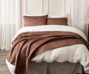 Bed with white linens and brown quilted throw pillows in front sunburst wall decor above sheer curtains framing the window