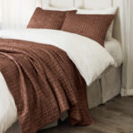 Bed with white sheets and a textured brown quilted throw and pillows laid along the headboard area
