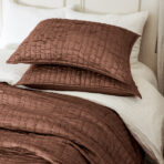 Brown quilted pillow on a bed with white pillows in the background close up view of textured fabric