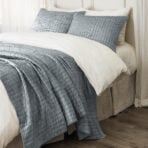 Bed with white linens and a textured blue quilted throw across the foot of the bed two matching blue pillows in front of white pillows