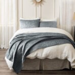 Bed neatly made with white sheets and a textured gray quilt in a bright bedroom with sheer curtains and a sunburst mirror above the headboard