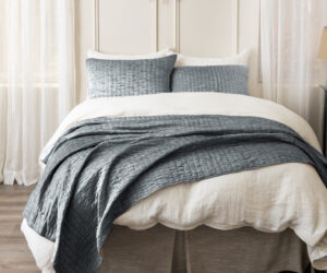 Bed neatly made with white sheets and a textured gray quilt in a bright bedroom with sheer curtains and a sunburst mirror above the headboard