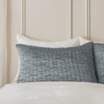 Bed setup with white linens and blue gray quilted pillows against a light beige paneled headboard
