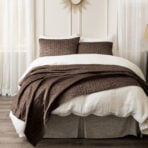 Bed with white sheets brown quilted blanket and matching pillows sheer curtains on both sides of a sunburst mirror above the headboard
