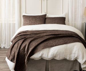 Bed with white sheets brown quilted blanket and matching pillows sheer curtains on both sides of a sunburst mirror above the headboard