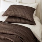 Brown quilted throw pillows stacked on a white bed with soft lighting in a neutral bedroom setting
