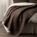 Close up of a brown quilted bedspread folded over a white sheet on a wooden floor