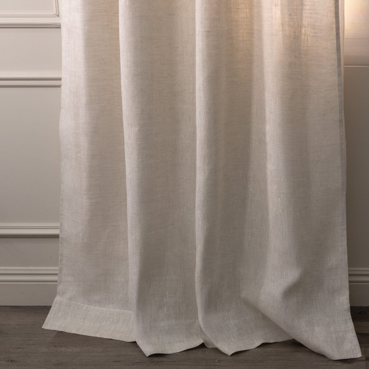 Beige curtain hanging in front of a wall with decorative molding and a wooden floor visible at the bottom edge