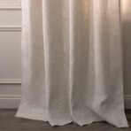 Beige curtain hanging in front of a wall with decorative molding and a wooden floor visible at the bottom edge