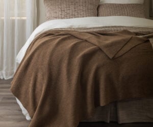 Bed with brown quilted pillows white sheets and a brown blanket in a softly lit bedroom Curtain to the left and wooden floor visible