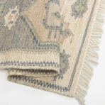Close up of a woven rug with blue and beige stripes and fringed edges on a white surface
