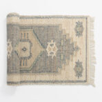 Corner of a beige and blue woven rug with a fringed edge on a white background showing geometric motifs and texture