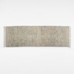 Partial view of a blue gray oriental rug with beige embroidery and fringed edges on a white background