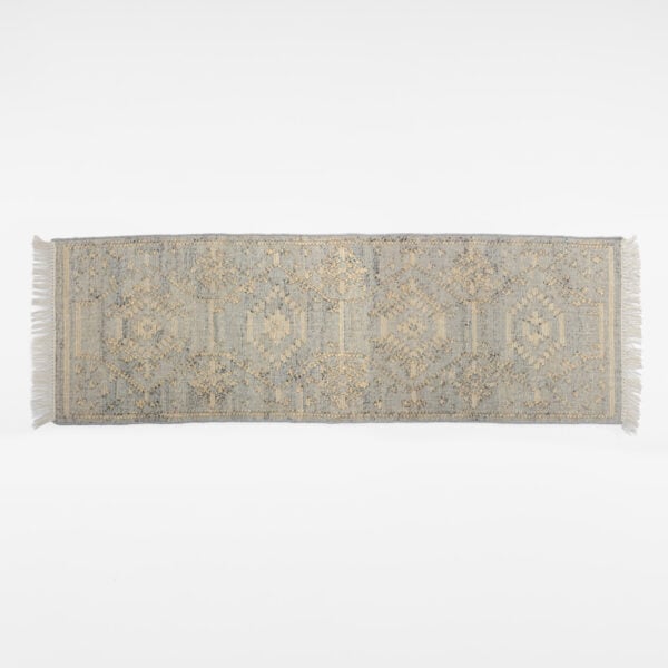 Partial view of a blue-gray oriental rug with beige embroidery and fringed edges on a white background