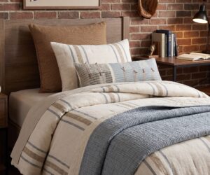 Bed with striped beige and blue quilt and tan pillows against a brick wall near a wooden headboard and nightstand lamp