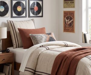 Bedroom corner with a bed beige and rust bedding and a wall of framed vinyl records retro posters on the right wall