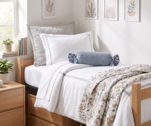 Minimalist bedroom scene with white bedding a gray quilted pillow a blue bolster and light wood furniture framed botanical art on a pale wall
