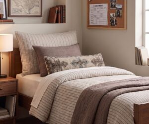 Cozy bedroom corner with a striped quilted bed two accent pillows a wooden nightstand and a corkboard on the wall with photos and notes
