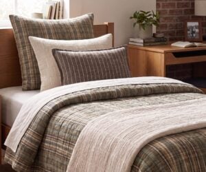 Cozy bedroom corner with a bed covered in plaid beige and gray bedding and pillows a wooden desk plant and brick wall in the background
