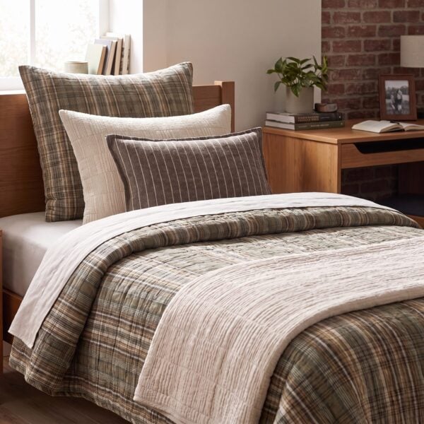 Cozy bedroom corner with a bed covered in plaid beige and gray bedding and pillows a wooden desk plant and brick wall in the background