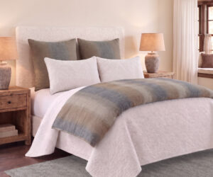 Hotel style bedroom with a white quilted bed gray pillows wooden nightstands and warm table lamps on each side Soft light filters through sheer window curtains to the right