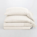 Stack of white pillows with a cream knitted blanket draped over the right edge on a pale background