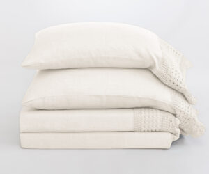 Stack of white pillows with a cream knitted blanket draped over the right edge on a pale background