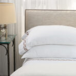 Close up of a neatly made bed with a beige upholstered headboard with nailhead trim a cream lampshade on a glass bedside table and sheer white curtains in the background