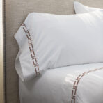 Close up of a white pillow with brown embroidered border resting on a beige upholstered headboardesque surface