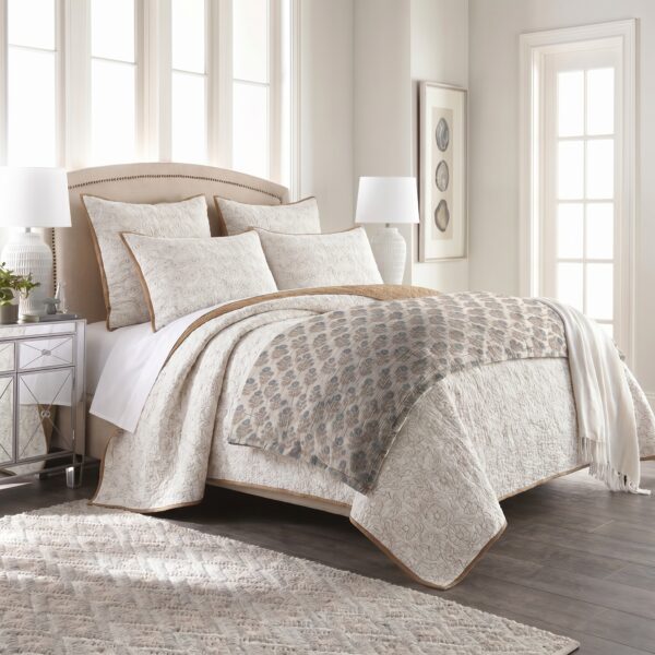Cream quilted bedding on a tufted headboard bed with decorative pillows in a bright, neutral bedroom with large windows.