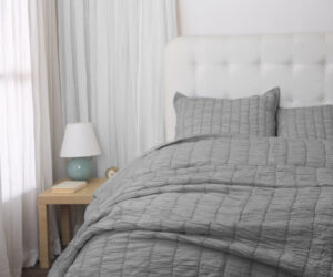 A neatly made bed with a light gray quilt and two matching pillows a white tufted headboard and a wooden nightstand with a lamp beside pale curtains