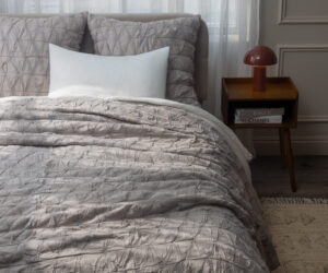 Queen bed with light gray quilted comforter and white pillow beside a wooden nightstand with a red lamp and sheer curtains in a neutral bedroom