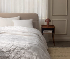 White quilted bed with pillows in a softly lit bedroom beige headboard sheer curtains and a wooden nightstand with a brown lamp on the right