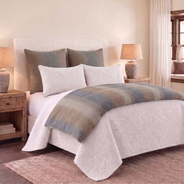 Bed with white quilted comforter and gray throw pillows in a neutral beige bedroom with two bedside lamps and wooden nightstands