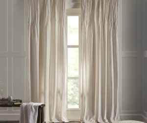 White flowing curtains framing a tall window in a sunlit bedroom with a wooden bench and cozy rug nearby