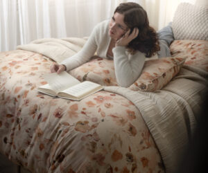 Woman lounging on a floral quilted bed propped on one elbow reading an open book