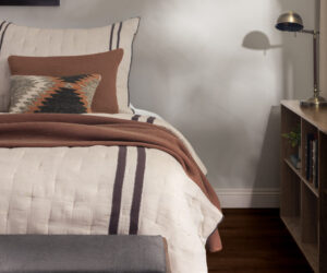 Bedroom scene with a white quilted bed rust colored pillows and a decorative zigzag pillow against a gray wall